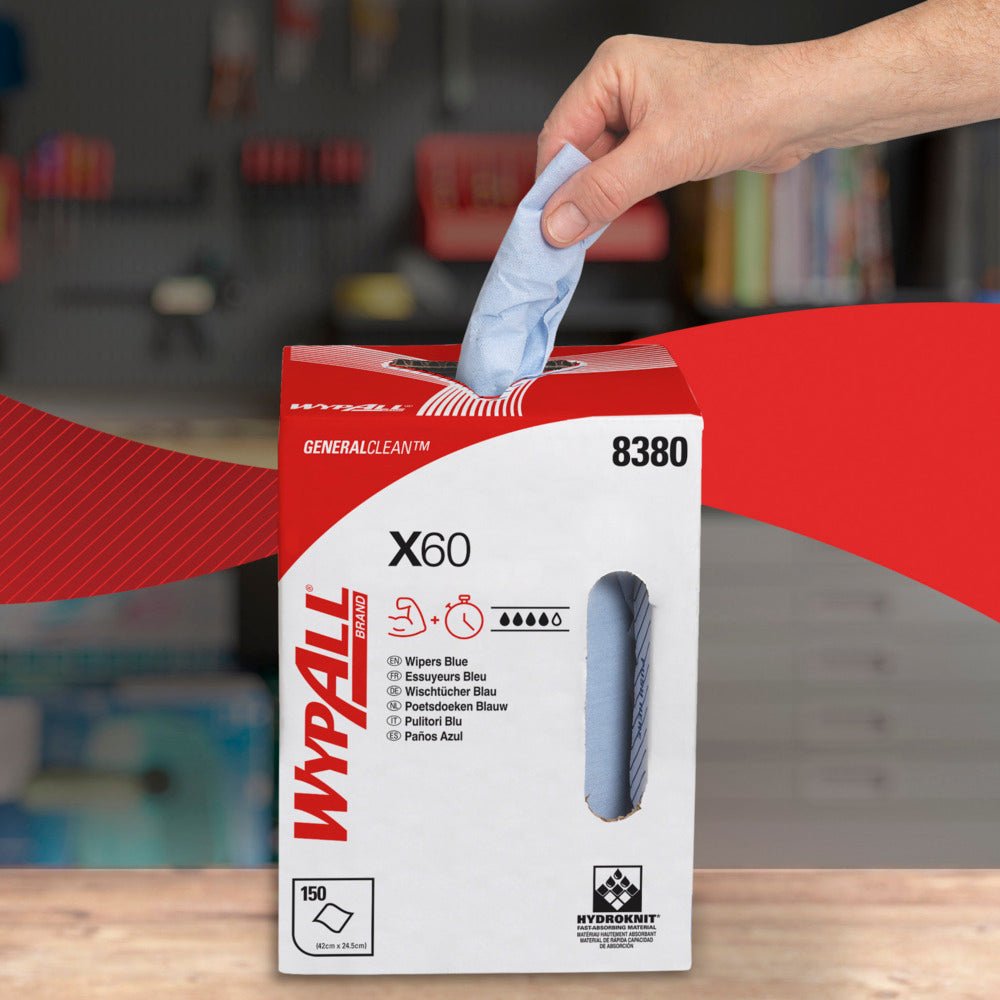 A hand pulls a blue WypAll® X60 General Clean sheet from a white-red dispenser box of Kimberly-Clark GmbH with the label "8380", "150 sheets", and "Central Dispensing". A blurred workshop with shelves is visible in the background.