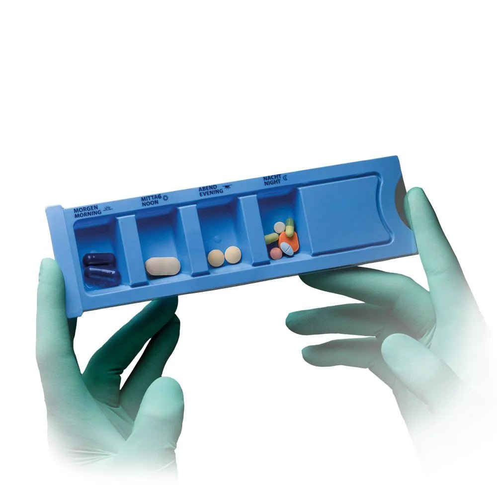 A person with green medical gloves holds a blue AMPri Med-Comfort PP disposable medication dispenser (4 compartments, 22.2 x 6 x 1.5 cm), with compartments for morning, noon, evening, and night.