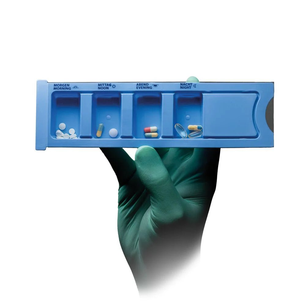 A gloved hand holds a blue Med-Comfort PP disposable medication dispenser (4 compartments, 22.2 x 6 x 1.5 cm) from AMPri Handelsgesellschaft mbH, each labeled compartment is filled with tablets; the background is white.