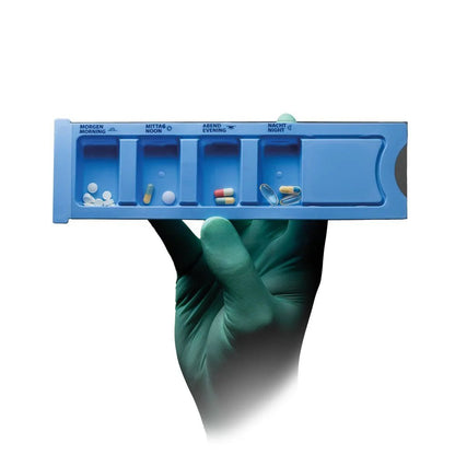 A gloved hand holds a blue Med-Comfort PP disposable medication dispenser (4 compartments, 22.2 x 6 x 1.5 cm) from AMPri Handelsgesellschaft mbH, each labeled compartment is filled with tablets; the background is white.