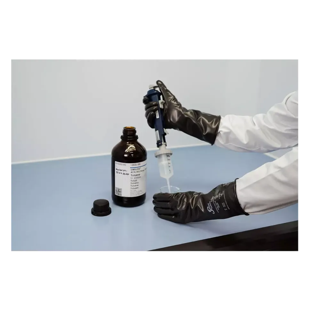 A person wearing AMPri SolidSafety ChemV Full Stable Brom-Butyl Chemical Protection Gloves with Viton outer layer and a white lab coat is using a pipette to draw liquid from a brown bottle on a light gray surface. The cap is nearby in the laboratory workspace, ensuring chemical resistance when handling substances.
