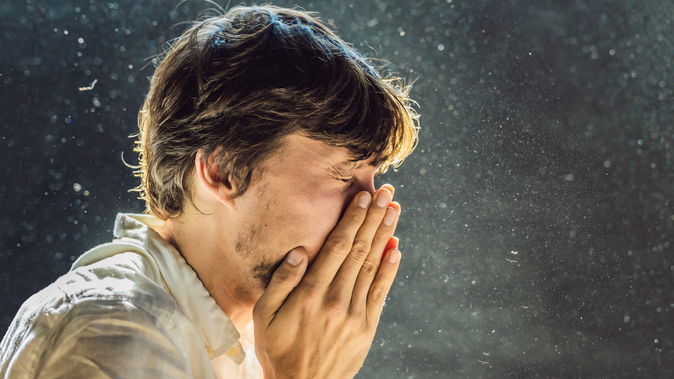 Eine Person niest mit geschlossenen Augen in die Hände, umgeben von sichtbaren Partikeln in der Luft und Staub vor einem dunklen Hintergrund.