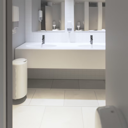 The public restroom presents itself in a modern elevation design with white tiled walls and floors. A long white countertop houses two sinks under a large mirror, as well as a Tork 553000 dispenser for zigzag towels Elevation H3 from TORK – Essity Professional Hygiene Germany GmbH, which offers efficient single sheet dispensing for paper towels. A conveniently positioned trash can is located near the entrance.