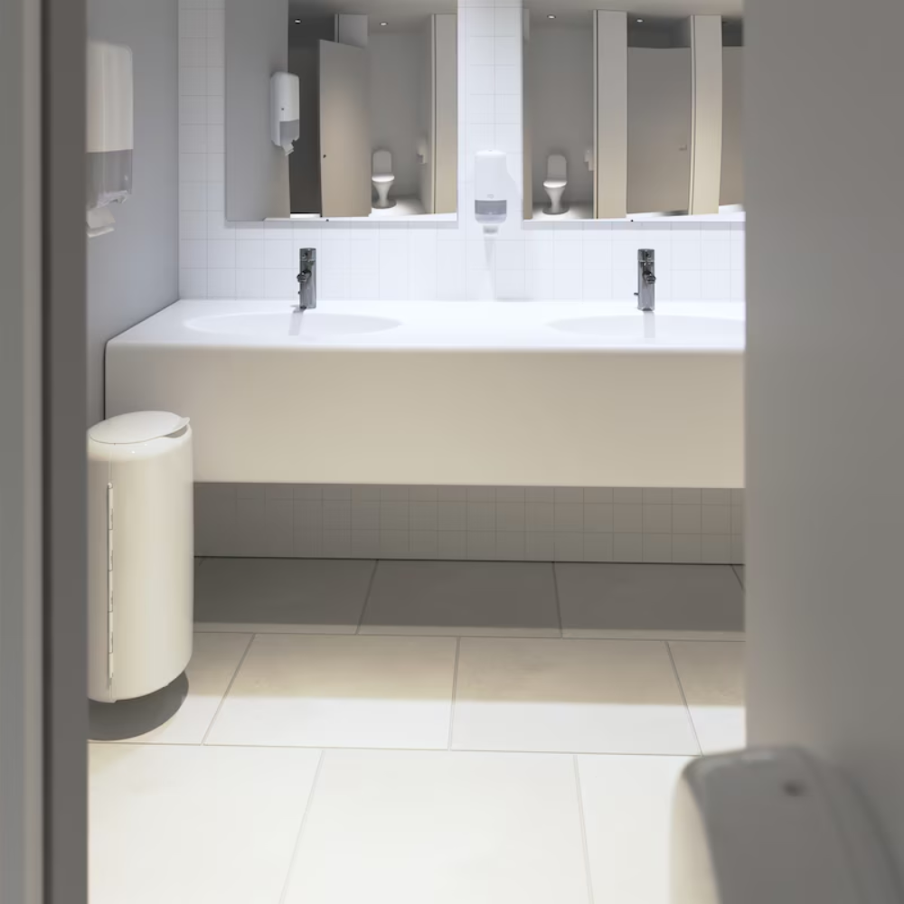 A clean, modern bathroom presents an Elevation design with a long white countertop featuring two sinks with chrome fixtures. The walls are adorned with white tiles and mirrors are mounted above the sinks. Tork 561500 dispensers for soap and hand sanitizer Elevation S4 by TORK – Essity Professional Hygiene Germany GmbH are elegantly mounted on the wall. An elegant white trash bin stands on the floor.