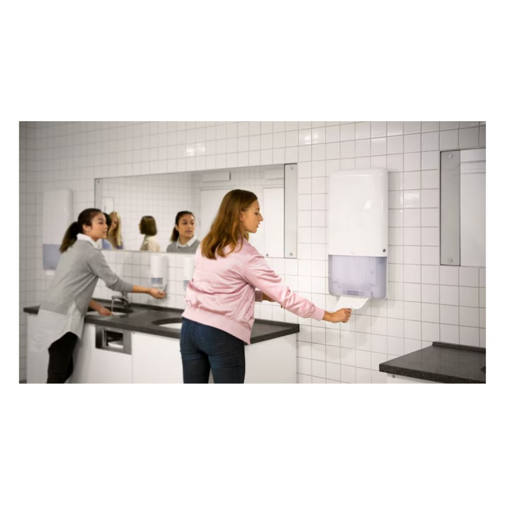 Two women stand in a bathroom with white tiled walls and large mirrors. One uses a sink on the left side while the other effortlessly pulls a continuous towel from a Tork PeakServe® 552500 dispenser for continuous towels Elevation H5 by TORK – Essity Professional Hygiene Germany GmbH, located on the right side. The gray countertop complements the bright lighting in this high-traffic washroom.