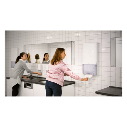 Two women stand in a bathroom with white tiled walls and large mirrors. One uses a sink on the left side while the other effortlessly pulls a continuous towel from a Tork PeakServe® 552500 dispenser for continuous towels Elevation H5 by TORK – Essity Professional Hygiene Germany GmbH, located on the right side. The gray countertop complements the bright lighting in this high-traffic washroom.