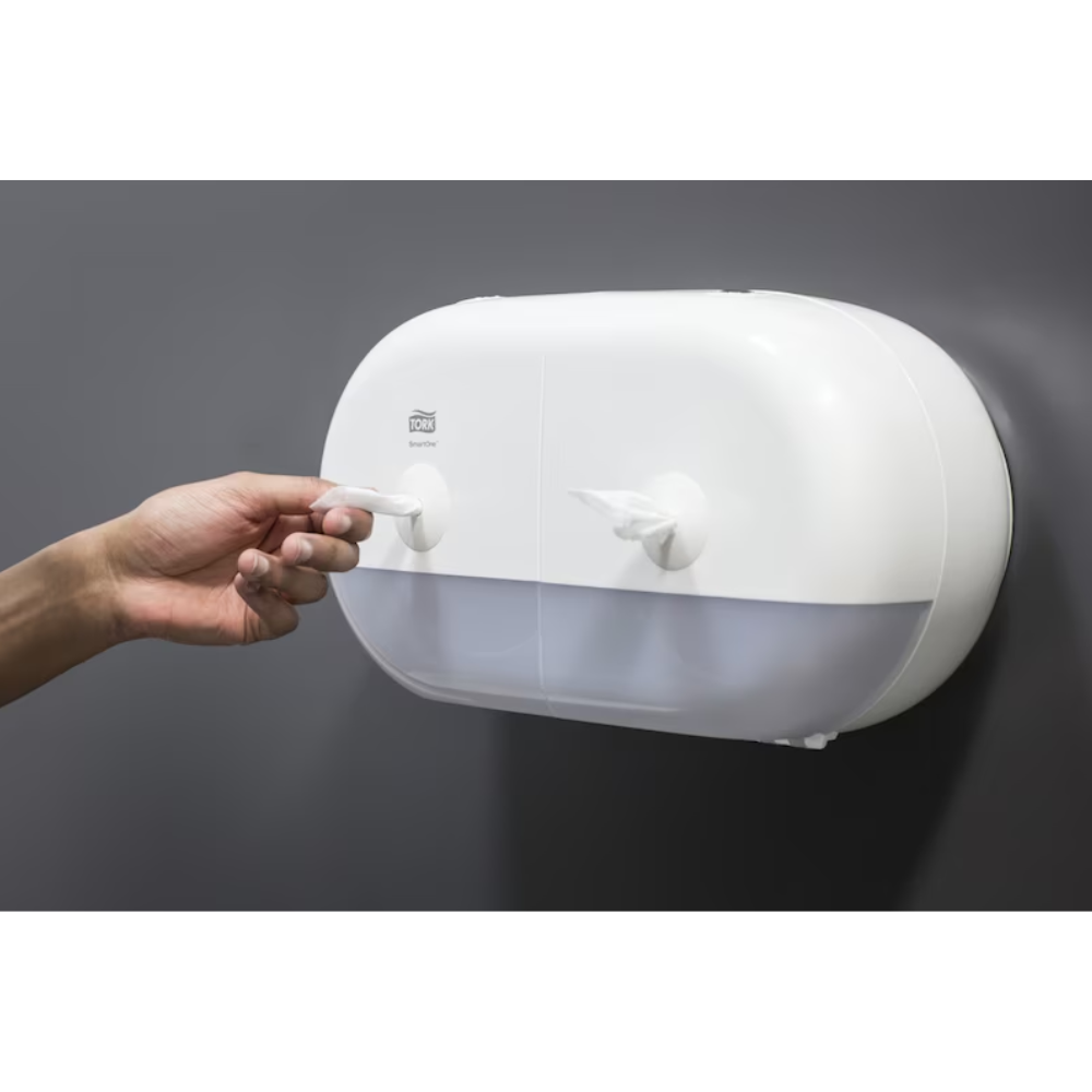 Eine Person zieht Papierhandtücher aus einem weißen, wandmontierten Tork SmartOne® 682000 Mini Doppelrollenspender für Toilettenpapier T9 | Packung (1 Stück). Der Spender hat eine gewölbte Oberseite und eine durchsichtige untere Frontplatte, auf der die Toilettenpapierrollen zu sehen sind. Der Hintergrund ist eine schlichte dunkle Wand.