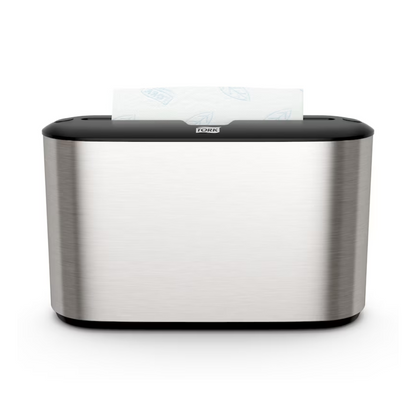 Pictured is the Tork Xpress® 460005 table dispenser for multifold towels Image Line H2 in stainless steel with a black top. From above, a multifold paper towel with a leaf motif is partially visible. On the front, the brand name “TORK – Essity Professional Hygiene Germany GmbH” is displayed, emphasizing compatibility with multifold towels.
