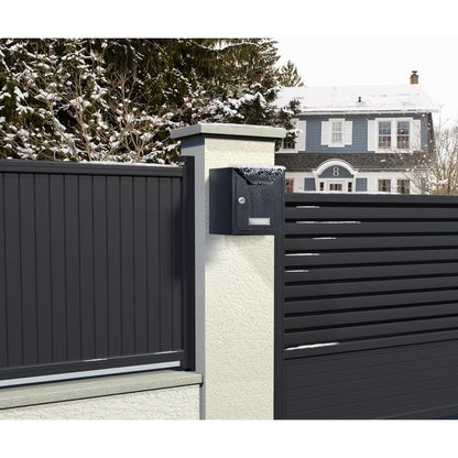 A modern black mailbox with snow is mounted on a white pillar next to a metal fence, secured with Scotch-FIX™ Extreme double-sided outdoor mounting tape from 3M Deutschland GmbH, with a blue-gray house and snowy trees in the background.