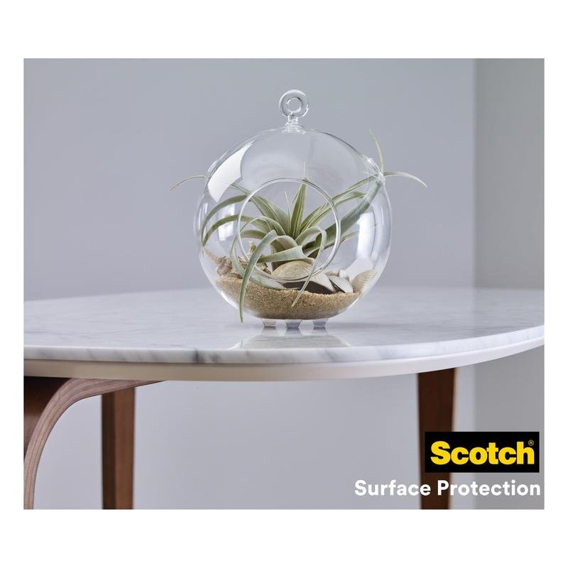A glass terrarium with air plants and sand stands on a white marble tabletop with wooden legs. In the corner of the table is the logo of 3M Germany GmbH and "Scotch® shock-absorbing elastic bumpers, self-adhesive, transparent."