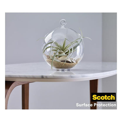 A glass terrarium with air plants and sand stands on a white marble tabletop with wooden legs. In the corner of the table is the logo of 3M Germany GmbH and "Scotch® shock-absorbing elastic bumpers, self-adhesive, transparent."