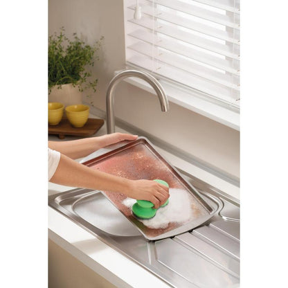 Someone scrubs a large, dirty baking sheet with the Scotch-Brite® Classic Cleaning Sponge from 3M Deutschland GmbH under a faucet in a bright kitchen with white blinds, a plant, and stacked yellow bowls in the background.