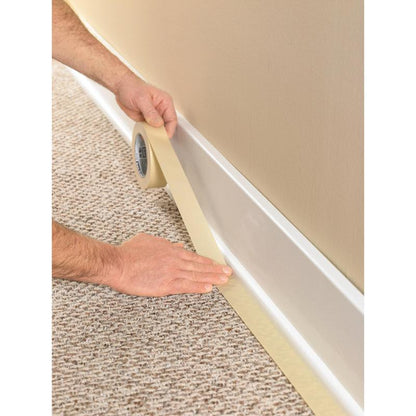 A person uses the Scotch® Classic Masking Tape (36 mm x 50 m, 1 roll/package, 70% PEFC) from 3M Deutschland GmbH to tape the edge of a white baseboard on light brown carpet before painting the beige wall above.