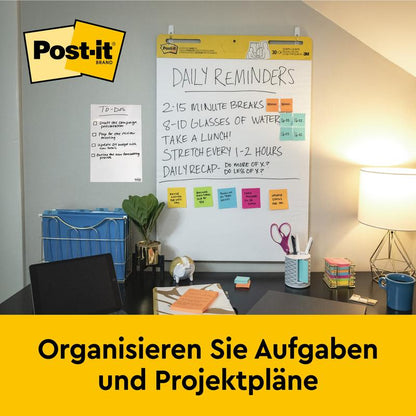 Ein Schreibtisch mit Büromaterial, Lampe und Stuhl steht vor einer an der Wand befestigten 3M Post-it® Super Sticky selbsthaftendes Meeting Chart (63,5x76,2 cm, 30 Blatt), das tägliche Erinnerungen und Notizen zeigt. Auf einem gelben Banner darunter steht "Organisieren Sie Aufgaben und Projektpläne.