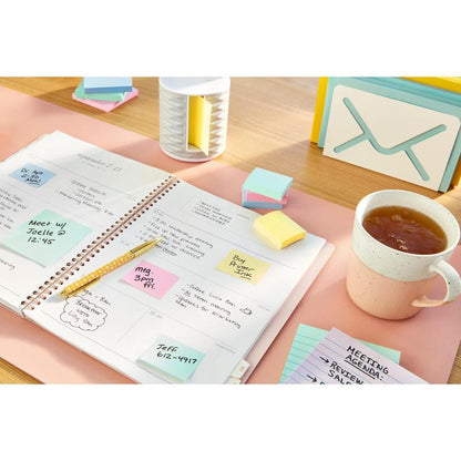 An open planner with handwritten notes, a pen, 3M Deutschland GmbH Post-it® Recycling Notes (76 mm x 127 mm, 100% PEFC), a cup of tea, an index card with the inscription "MEETING AGENDA" and desk accessories on a light pink desk pad.