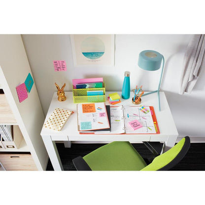 A white desk with an open folder, notebooks, 3M Post-it® Notes (76x76 mm, 100 sheets, 100% PEFC), writing materials, a lamp, a water bottle, and organizers. In front is a green chair; a picture and Post-it® Notes hang on the wall.