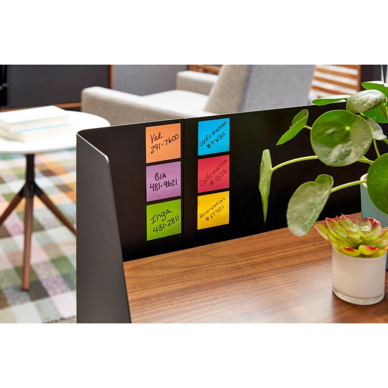 On a wooden desk are five colorful Post-it® Super Sticky Z-Notes (76 x 76 mm, 90 sheets/block) by 3M Deutschland GmbH. Next to them are a potted plant and a succulent. In the background: table, chairs, checkered carpet.