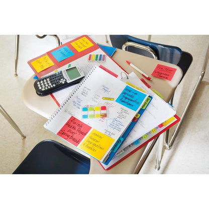 A student desk with notebooks, a calculator, pens, folders, and 3M Deutschland GmbH Post-it® Super Sticky Notes (47.6mm x 47.6mm, 90 sheets/block, 100% PEFC) with handwritten study notes; chairs are visible around the desk.